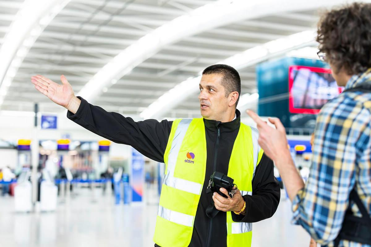 Male security guard at Heathrow Airport, wearing a Mitie branded high vis, pointing to the right and directing a customer