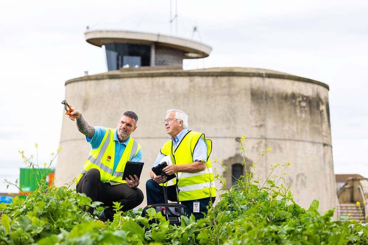Two male Mitie engineers wearing high vis vests, crouching in front of Martello Tower in Essex. The man on the right-hand side is pointing off-screen with a tool in his hand